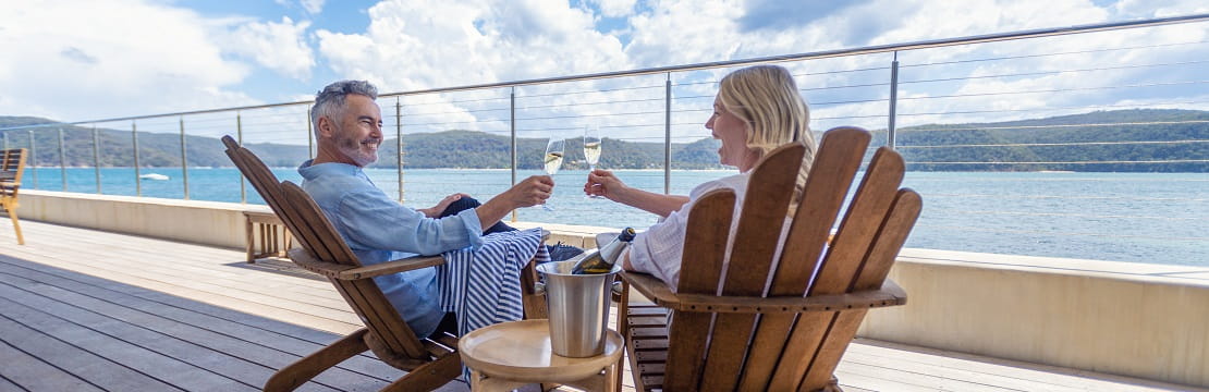A couple sits in chairs on a picturesque deck and toasts wine glasses. 